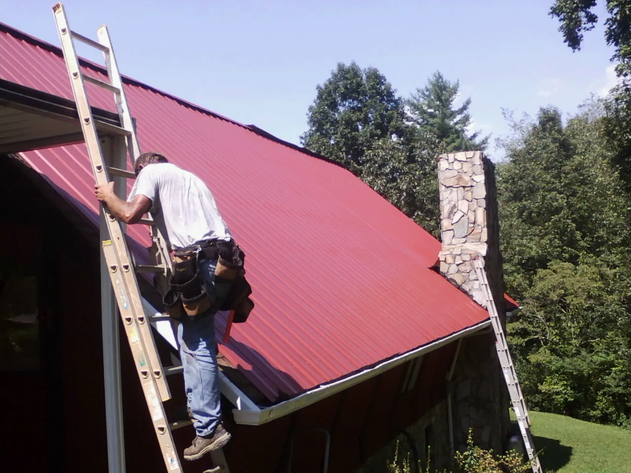 Roofer on a ladder beside a red metal roof and stone chimney — Soffit & Fascia Repair by Roofing Brothers in Ephrata